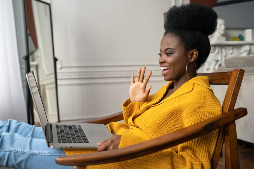 Smiling African American millennial woman sitting in chair talking in video chat on laptop say hello