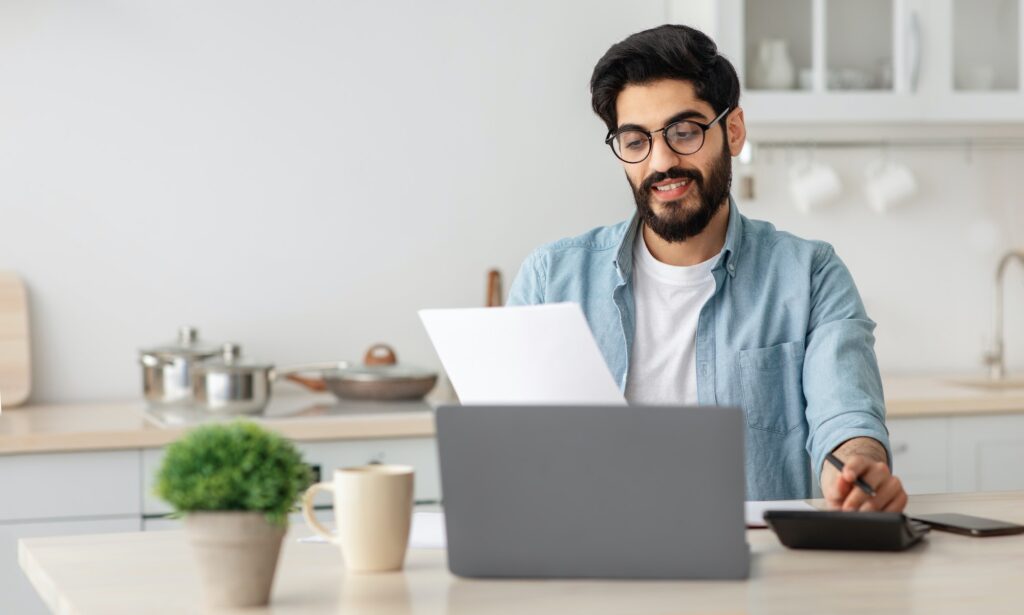 Portrait of happy arab guy reading insurance documents at home, sitting at table in kitchen and