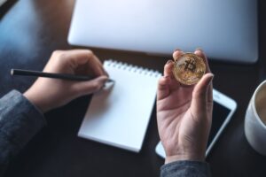 a woman's hands holding bitcoin and writing on empty notebook with laptop and mobile phone