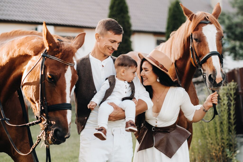 A family in white clothes with their son stand near two beautiful horses in nature. A stylish couple